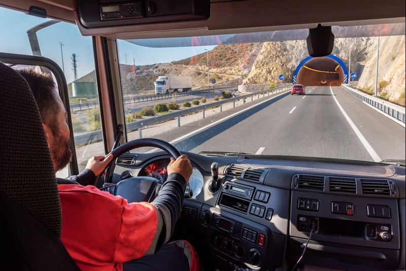 Un chauffeur de camion, vu de l'intérieur de la cabine de son camion, est assis derrière le volant. Il roule sur une autoroute. Son camion s'approche de l'entrée d'un tunnel.